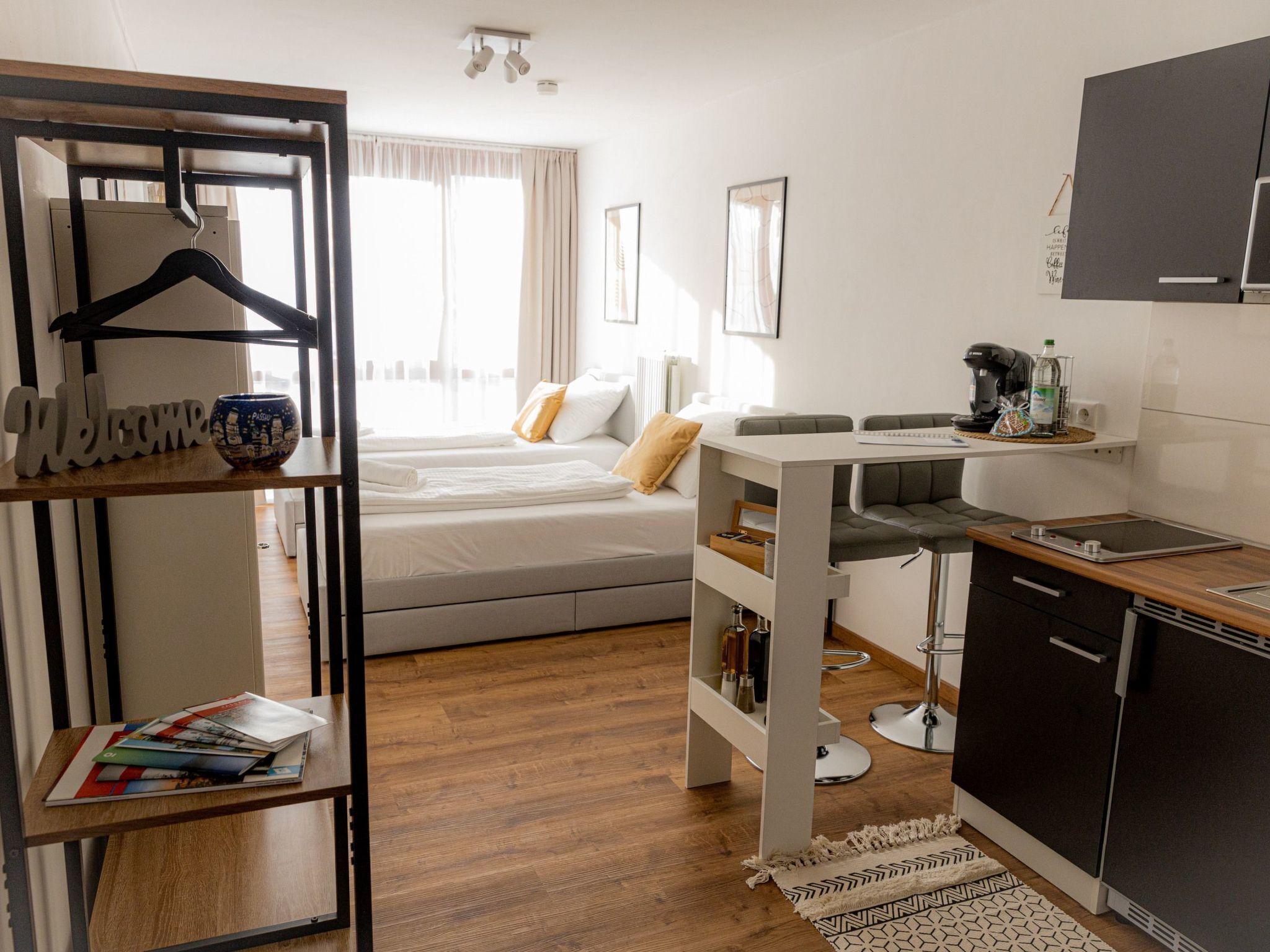 Une chambre d’hôtel compacte comprenant un lit, une petite kitchenette, une table en bois et une étagère sur laquelle est posée une pancarte « Bienvenue ».