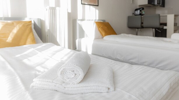 Quarto de hotel com duas camas, roupa de cama branca e uma toalha enrolada na cama.