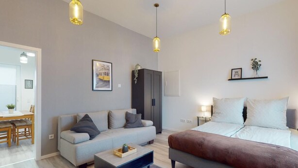 Un salon moderne comprenant un canapé, un lit et un coin repas. On y trouve des luminaires suspendus, un tableau encadré au mur et une petite table avec une plante.