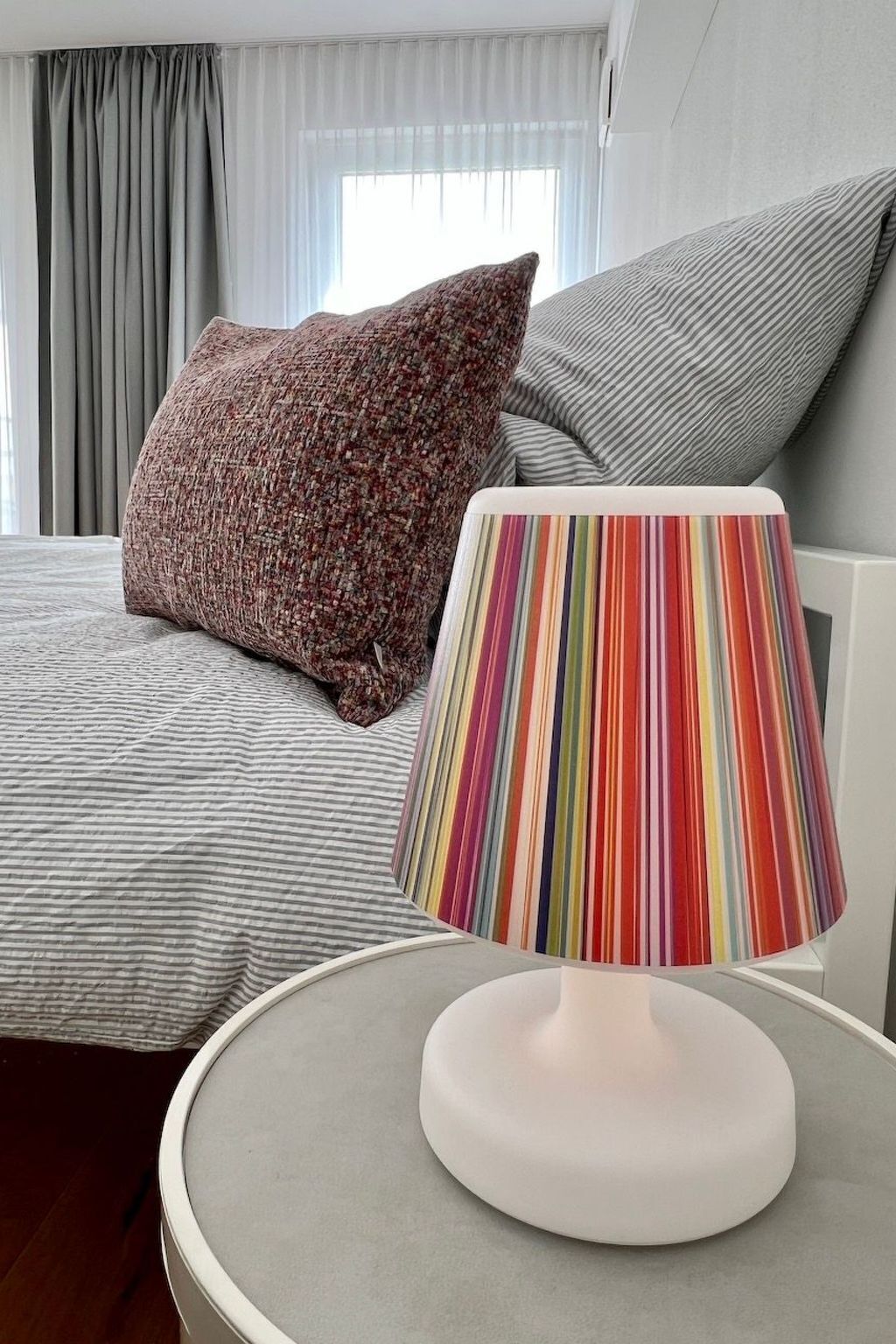 A bed with striped bedding, a round table with a colorful striped lamp, and a gray pillow.