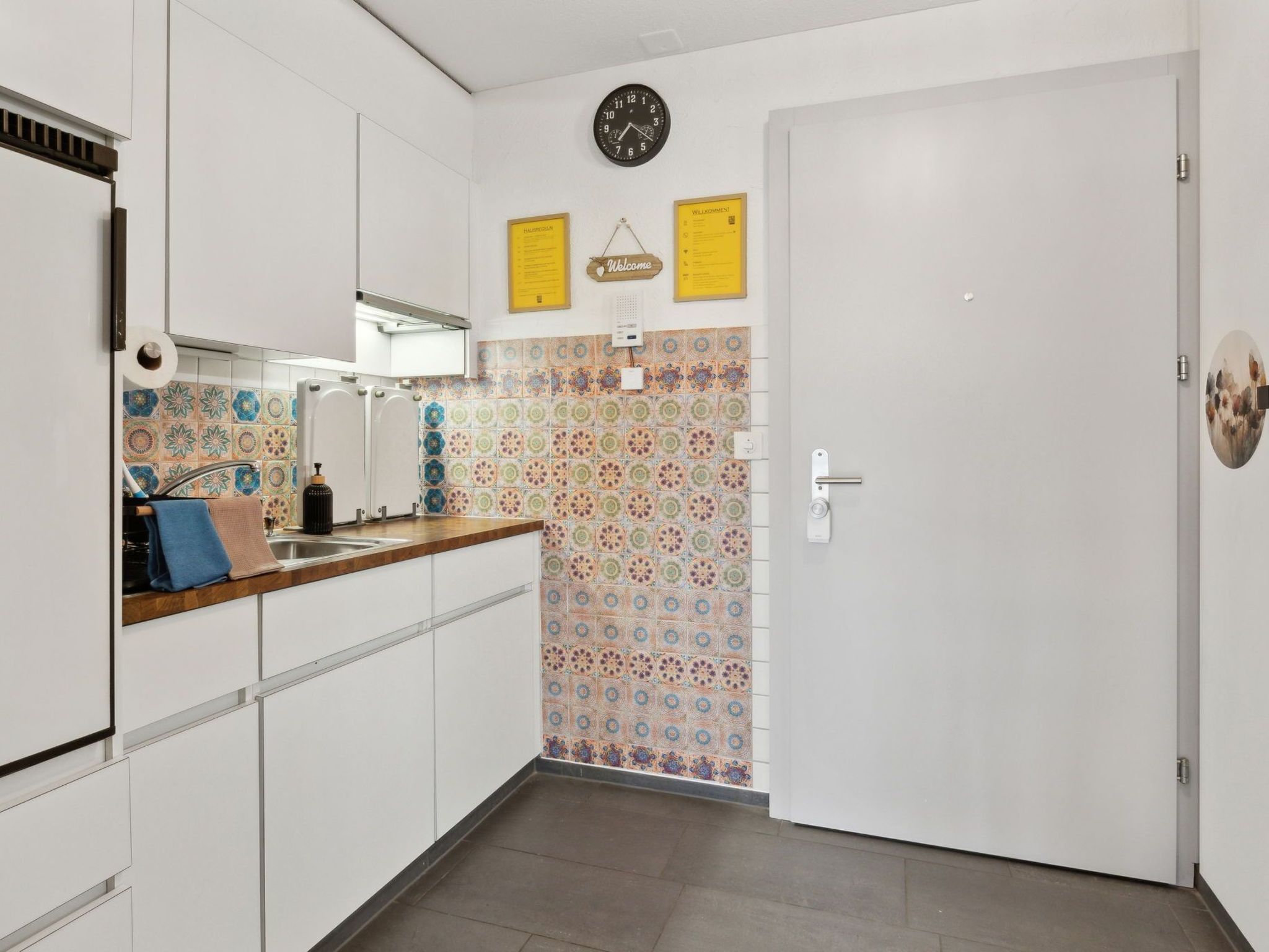 Cocina con gabinetes blancos, encimera de madera, un lavamanos y un salpicadero con diseño. Hay un reloj en la pared y una puerta con pomo.