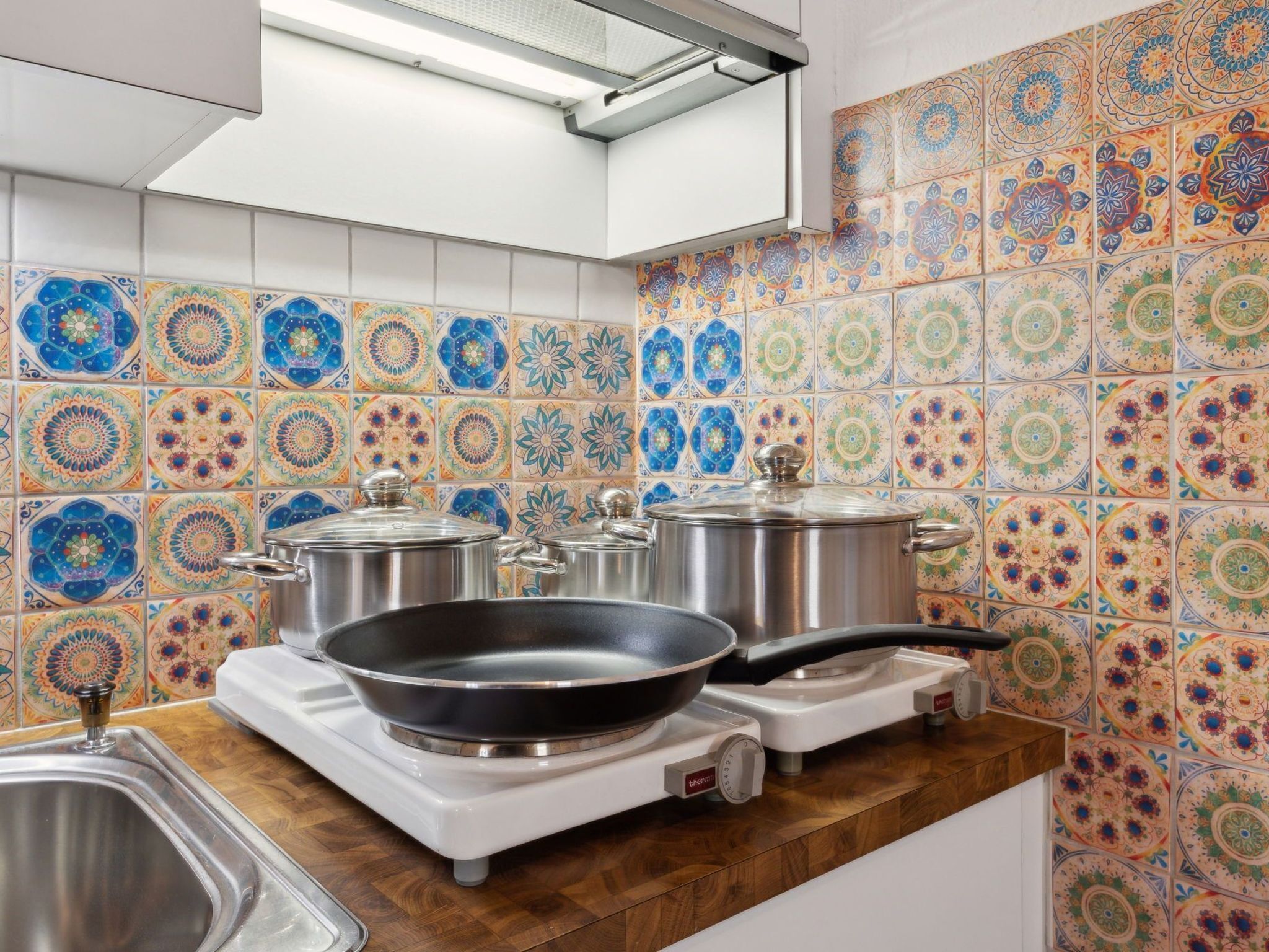 Une cuisine avec un dosseret de carrelage aux couleurs vives, des casseroles en acier inoxydable, une cuisinière à gaz et un évier.