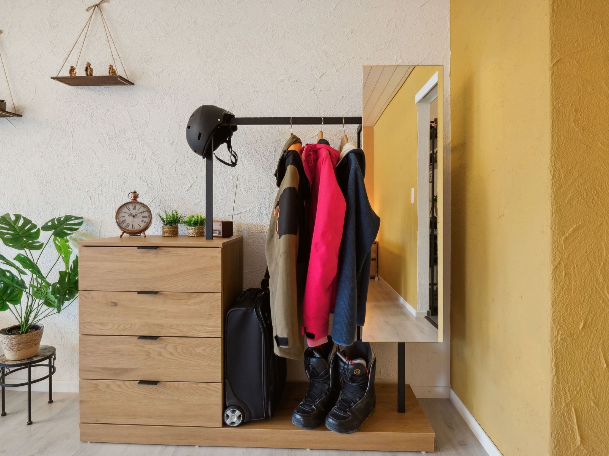 A compact room with a wooden dresser, a hanging coat rack, a helmet, a suitcase, and a small clock.