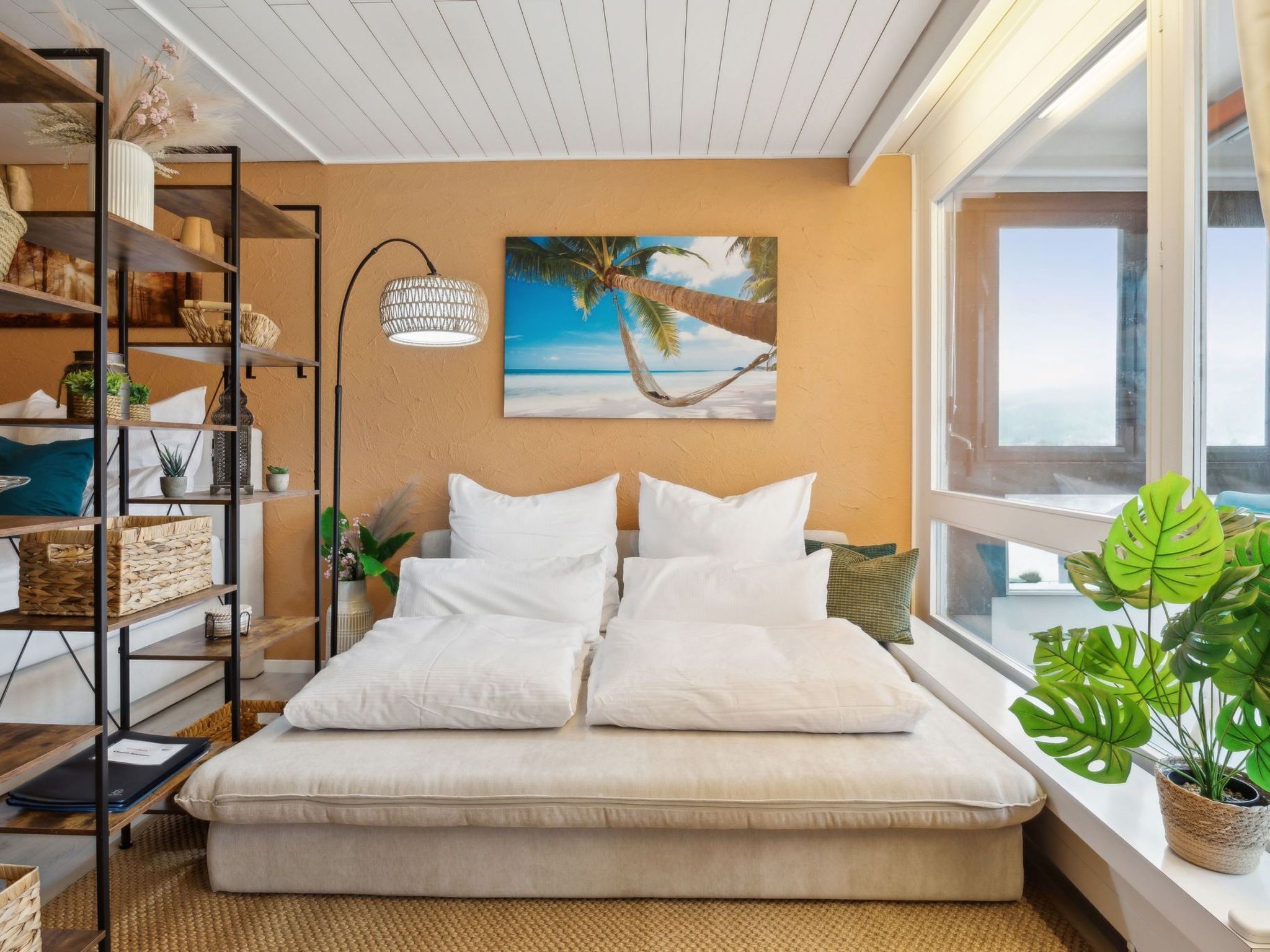 Une chambre à coucher avec un lit, une étagère ornée d’objets décoratifs, un tableau représentant une scène de plage et une fenêtre donnant sur un paysage agréable.
