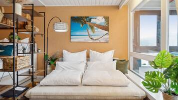 Une chambre à coucher avec un lit, une étagère ornée d’objets décoratifs, un tableau représentant une scène de plage et une fenêtre donnant sur un paysage agréable.
