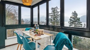 A room with a large window overlooking a lake and trees, a table with chairs, and a hanging light fixture.