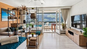 A modern living room with a sofa, coffee table, and TV. There is a balcony with a view of the sea.