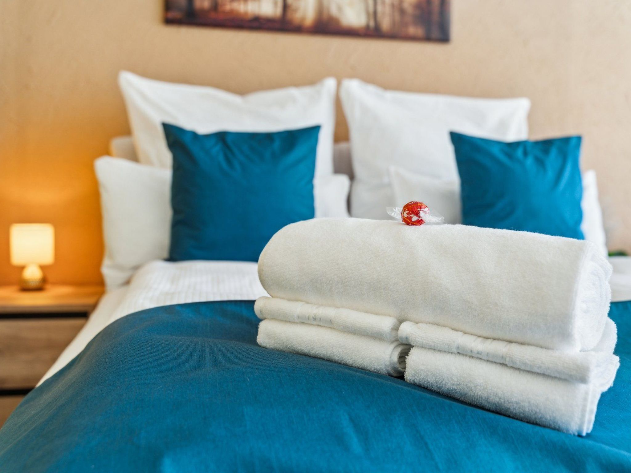 A neatly made bed with white and blue bedding, a red decorative item on the pillows, and a lamp on the nightstand.