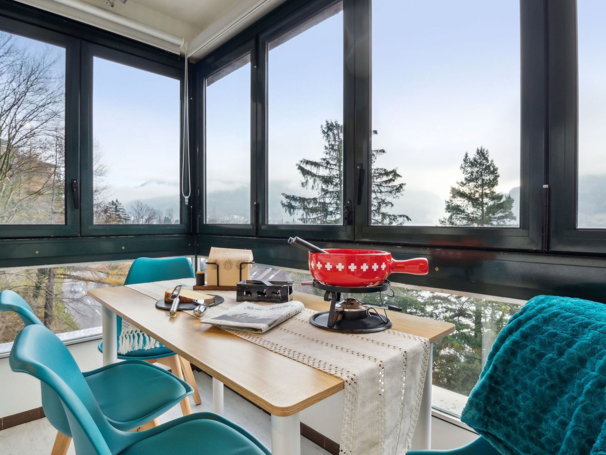 Een gezellige kamer met een eettafel, een rode pan op een fornuis, en uitzicht op bomen en bergen door grote ramen.