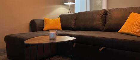 A modern living room with a sectional sofa, two orange cushions, a small round table with a candle, and a floor lamp.
