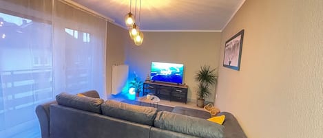 A living room with a large grey sofa, a flat-screen TV, a ceiling light fixture, and a potted plant.