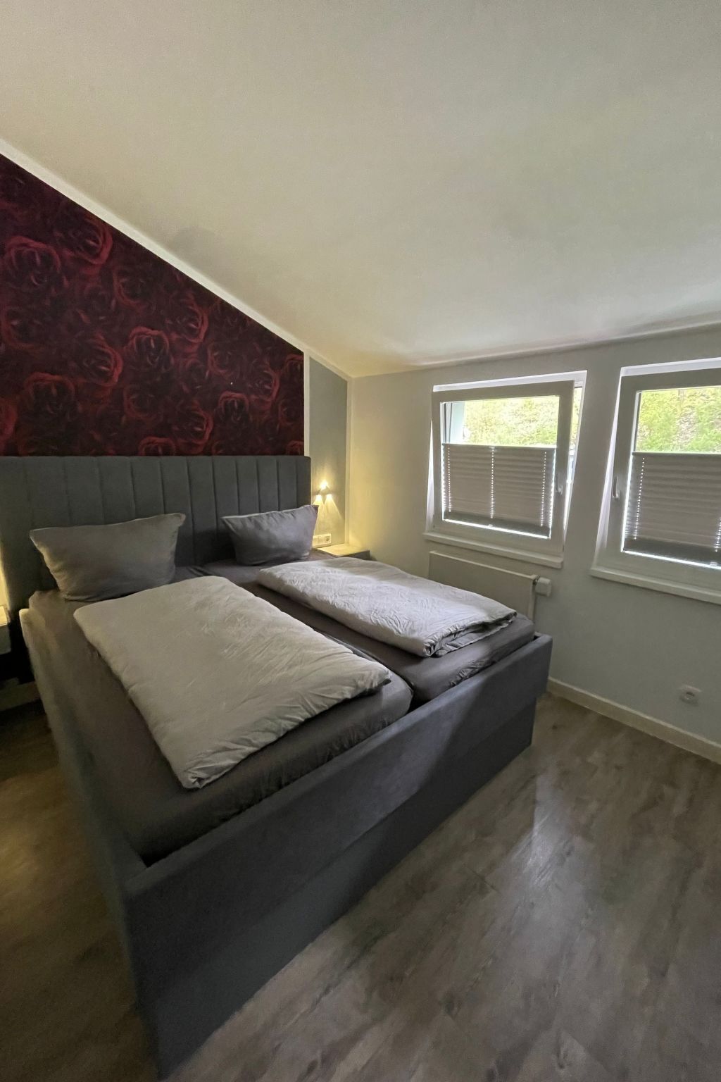 Une chambre avec un grand lit, deux tables de chevet, un mur décoré d’un motif rouge et deux fenêtres équipées de stores.