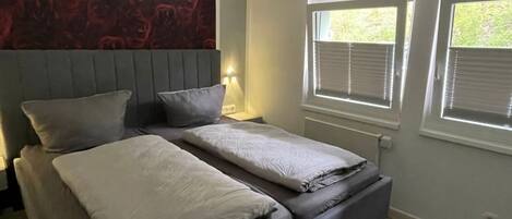 Une chambre avec un grand lit, deux tables de chevet, un mur décoré d’un motif rouge et deux fenêtres équipées de stores.