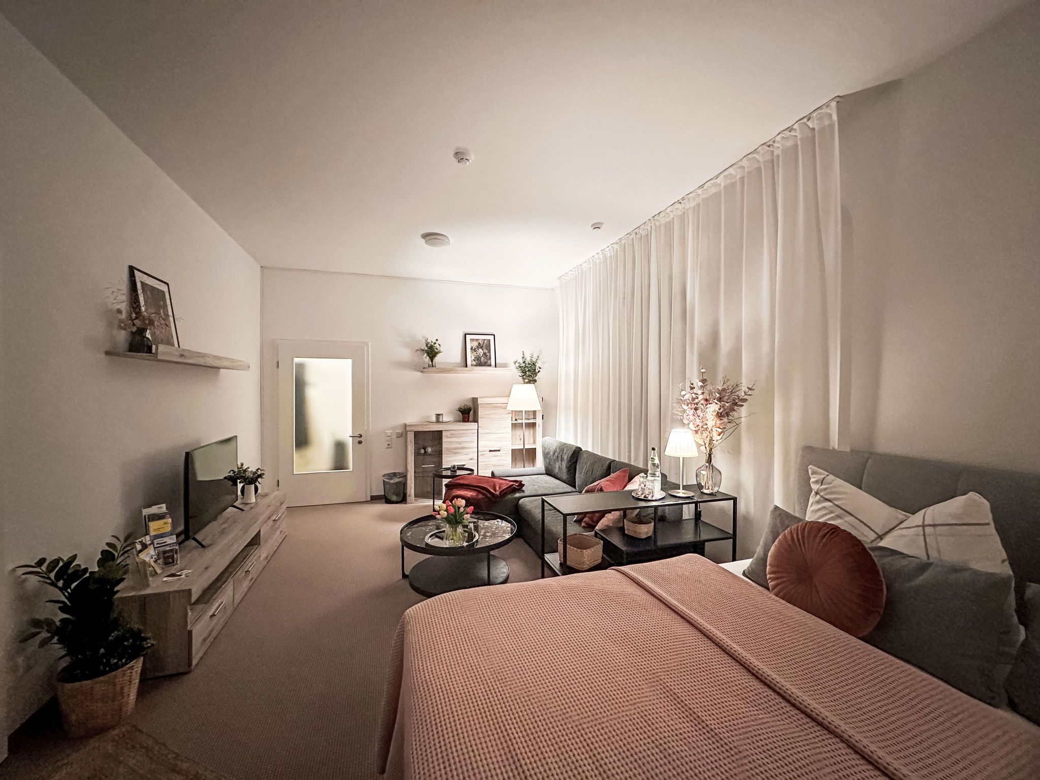 Un salon moderne avec un canapé, une table basse et un téléviseur. On y trouve un lit avec une couverture et des oreillers, ainsi qu’une petite table avec une lampe et des fleurs.