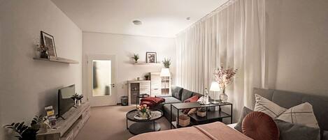Ein modernes Wohnzimmer mit einer Couch, einem Couchtisch und einem Fernseher. Es gibt ein Bett mit Decke und Kissen sowie einen kleinen Tisch mit einer Lampe und Blumen.