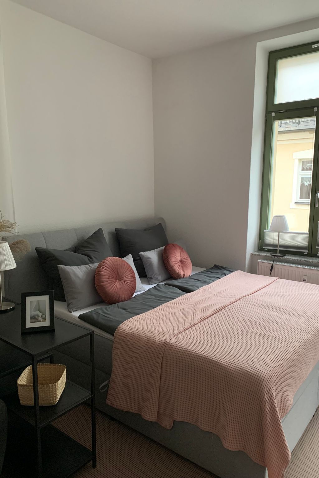 A bedroom with a bed, a bedside table with a lamp, a picture frame, and a window with blinds.