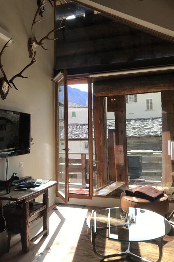 A room with a TV, a wooden table, a glass table, and a view of a snowy landscape through large glass doors.