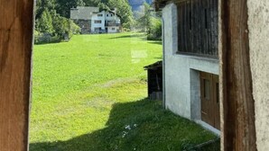 Un edificio rústico con fachada blanca y exterior de madera, frente a un paisaje de campos verdes y montañas.