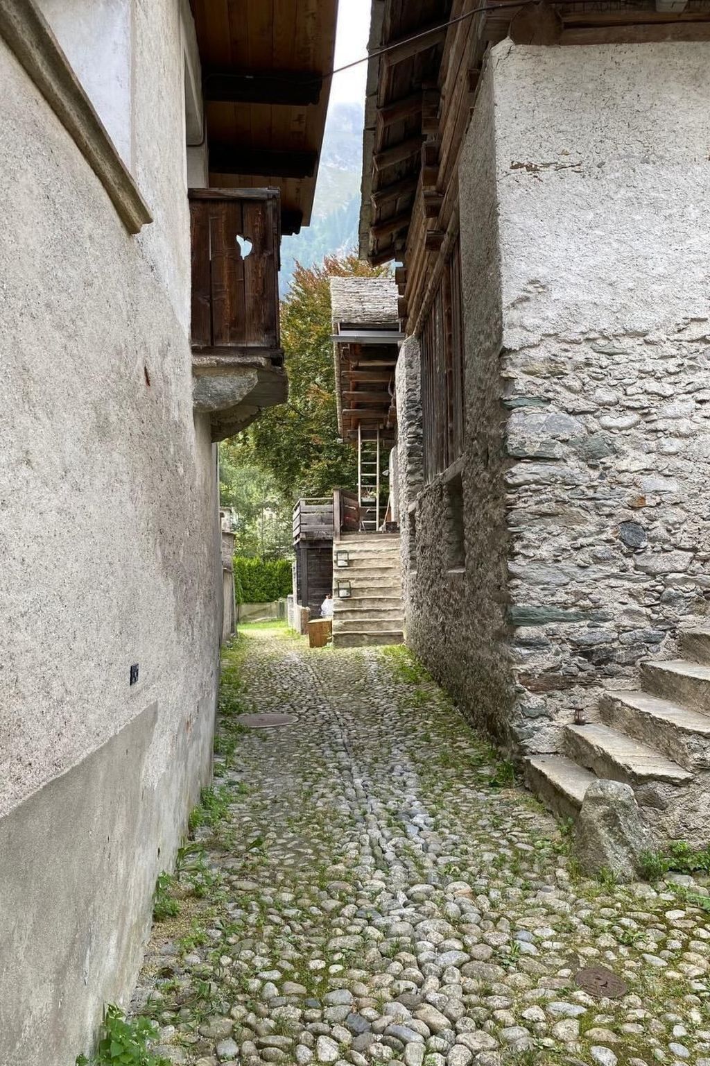 Un estrecho callejón adoquinado entre edificios de piedra con balcones de madera y una escalera de piedra que lleva a una puerta.