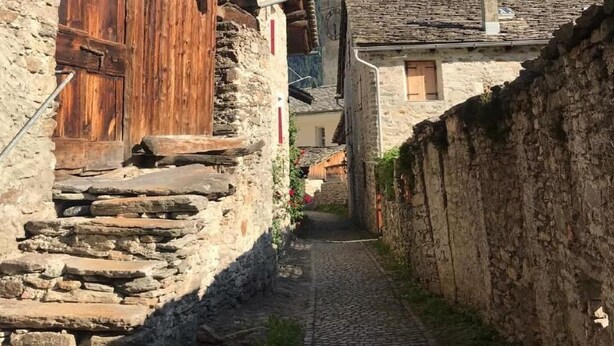 Una estrecha calle adoquinada bordeada por edificios de piedra rústica con puertas de madera y una torre de campanas al fondo.