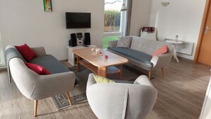 Ein modernes Wohnzimmer mit einer Couch, einem Sessel und einem Couchtisch. Es gibt einen Flachbildfernseher, Lautsprecher und ein Fenster mit Blick auf Grünflächen.