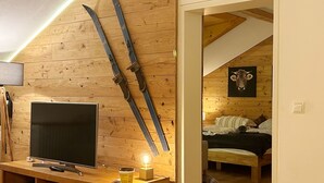 A wooden room with a TV on a wooden stand, a pair of skis leaning against the wall, and a bed with a cow picture on the wall. - FeWO Allgäu Chalet - gemütliche Hüttenatmosphäre und Schwimmbad und Sauna im Ha by Interhome (Oberstdorf)