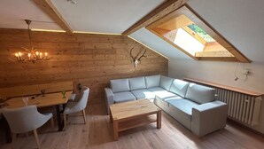 A cozy loft bedroom with a sloped ceiling, wooden walls, a large window, a sofa, a dining table, and a chandelier. - FeWO Allgäu Chalet - gemütliche Hüttenatmosphäre und Schwimmbad und Sauna im Ha by Interhome (Oberstdorf)