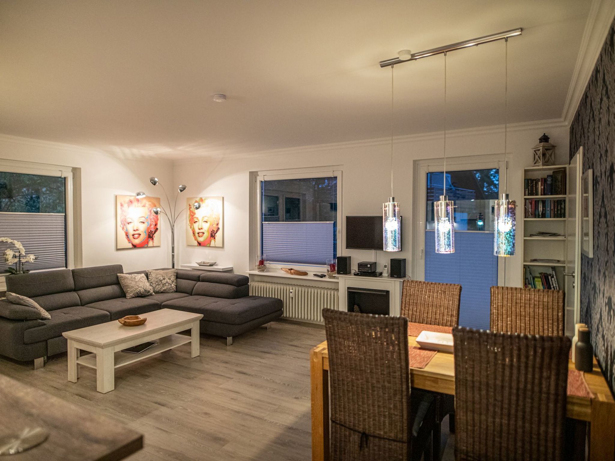 Een moderne woonkamer met een grijze bank, een witte salontafel en rieten fauteuils. Er is een televisie, een boekenkast en kunst aan de muren.