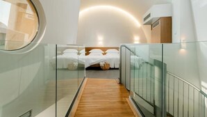 A modern hotel room with a glass-walled bed, a wooden floor, and a curved ceiling light.