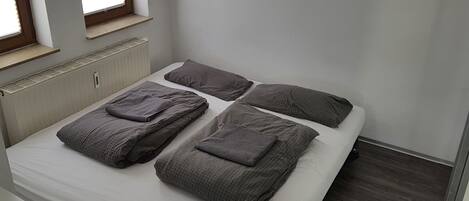 A single bed with four pillows, a bedside table, and a window with blinds.
