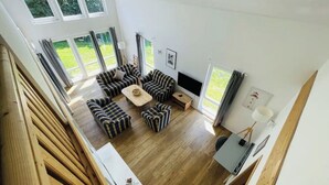 En rummelig stue med en stor sofasæt, et sofabord og et fladskærms-tv. Store vinduer giver naturligt lys og udsigt til det fri.