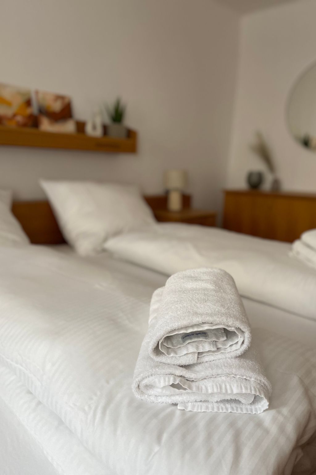 A neatly made bed with a folded towel on it, a bedside table with a lamp, and a shelf with decorative items.