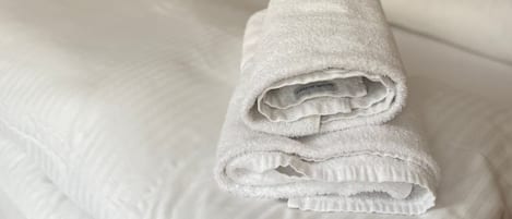 A neatly made bed with a folded towel on it, a bedside table with a lamp, and a shelf with decorative items.