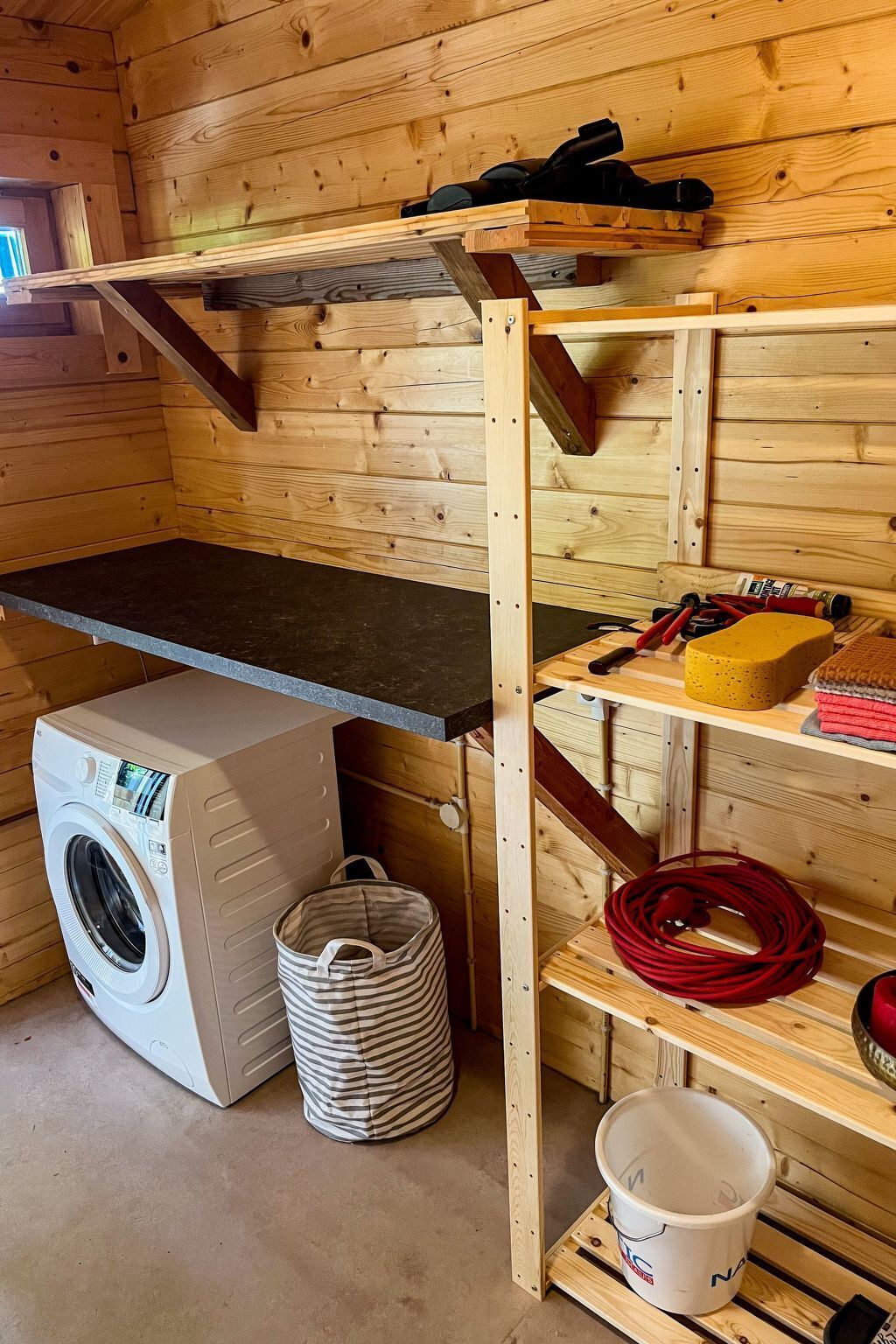 A laundry room with a washing machine, a drying rack, shelves with cleaning supplies, and a wooden bench.
