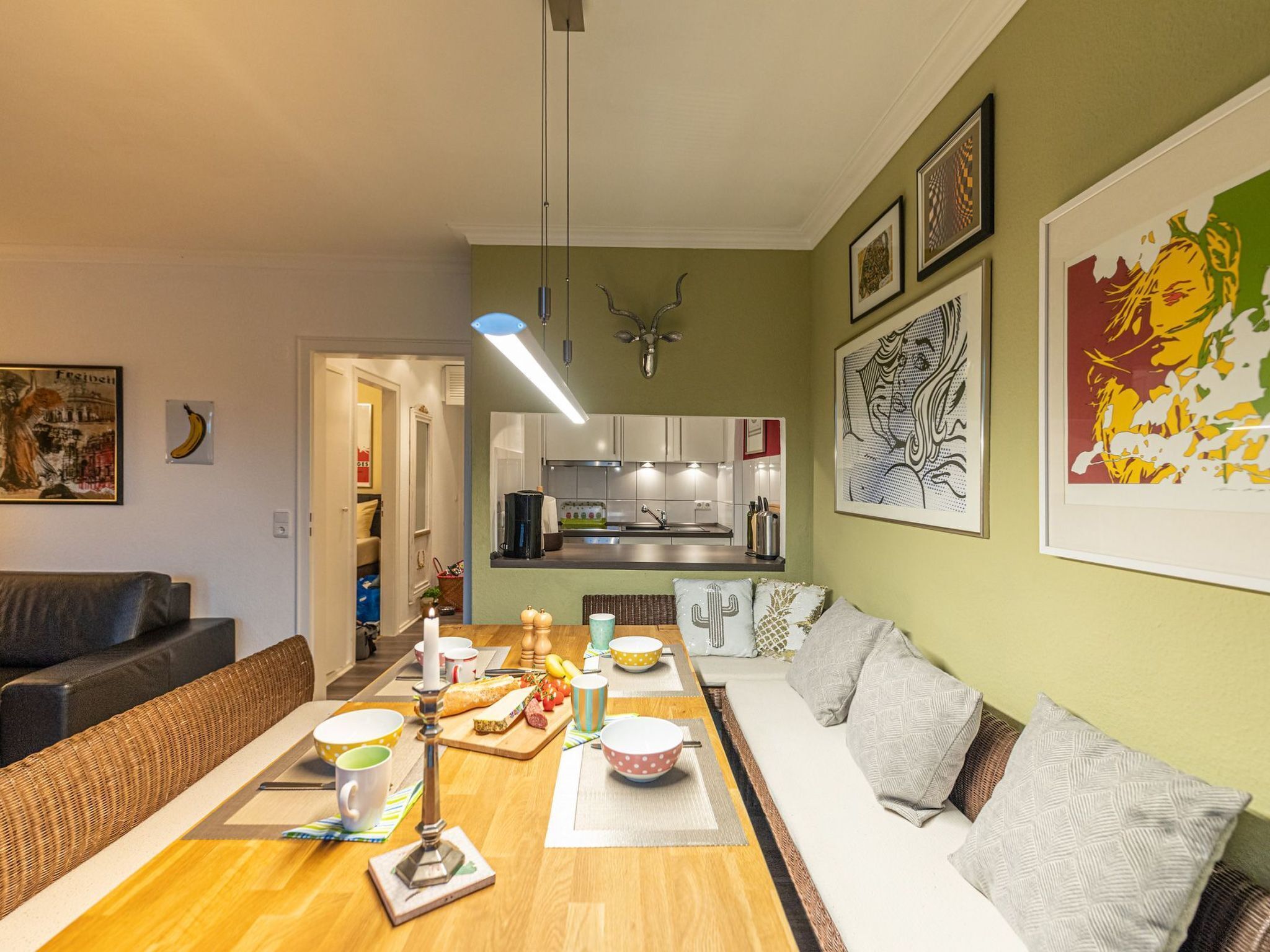 A modern kitchen with white cabinets, a dining area with a wooden table set for a meal, and a living room with a sofa and cushions.