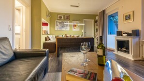 Un salon chaleureux avec un canapé en cuir, une table basse en bois et une cheminée. On y trouve également un coin repas avec une table et des chaises en osier, et une cuisine à l’arrière-plan.