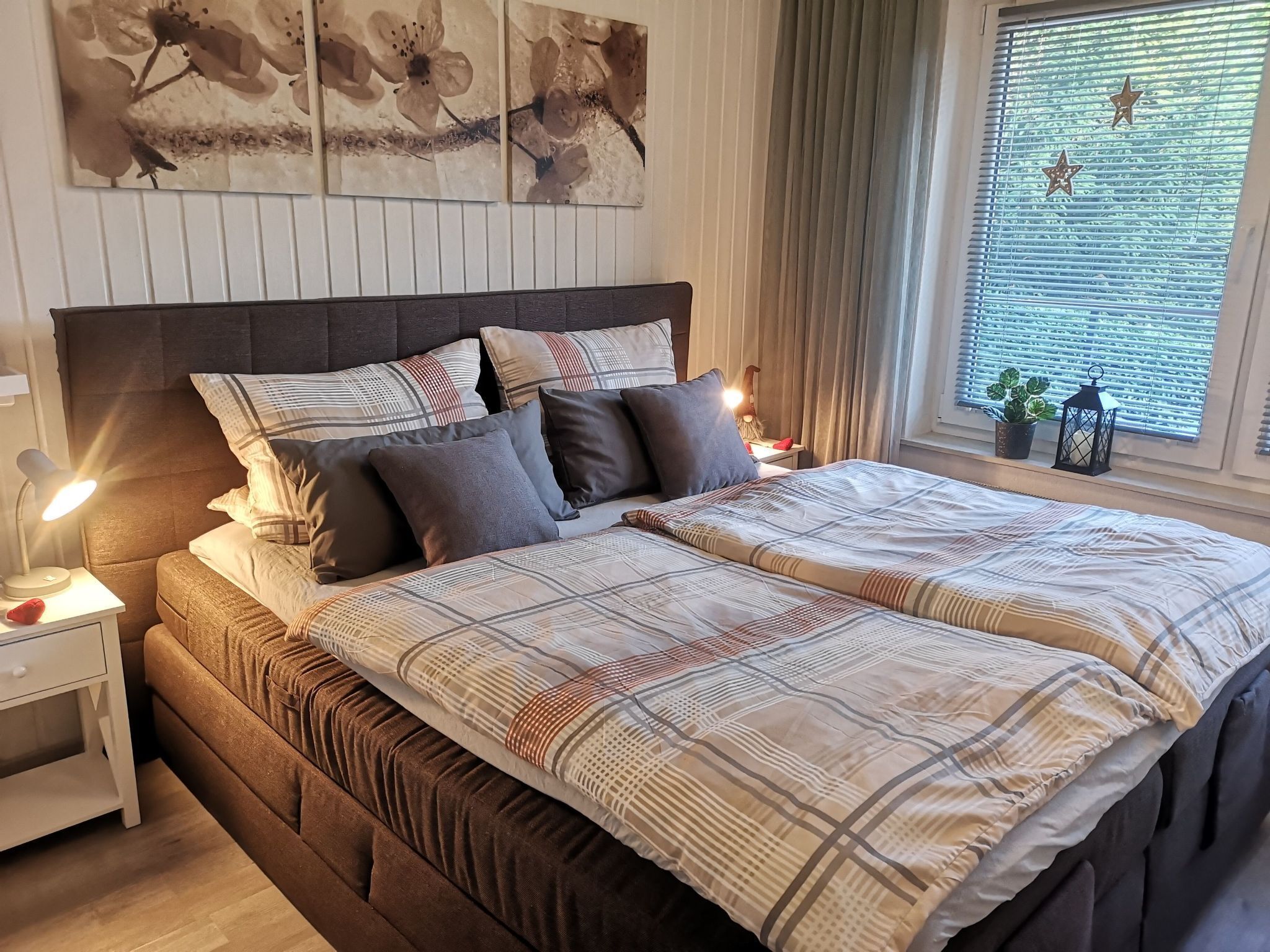 Ein Schlafzimmer mit einem großen Bett, vielen Kissen, einem Nachttisch mit Lampe, einem Fenster mit Jalousien und Wandbildern.