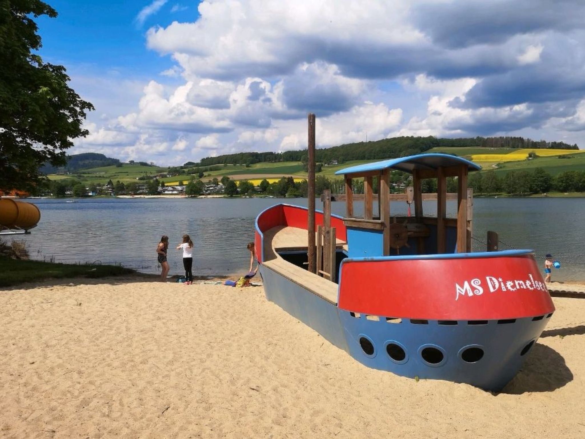 Ein kleines Boot mit Kabine und rot-blauer Farbgebung liegt an einem Sandstrand. Im Hintergrund sind ein See und Hügel zu sehen.