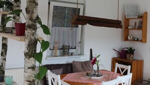 Un coin repas avec une table ronde, des chaises blanches et un banc en bois. On y trouve des étagères avec des plantes et une fenêtre donnant sur un beau paysage.