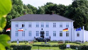 A large, white building with a tiled roof, multiple flags, and a well-maintained garden in front. - Villa Specht- Ferienbungalow by Interhome (Neubukow)