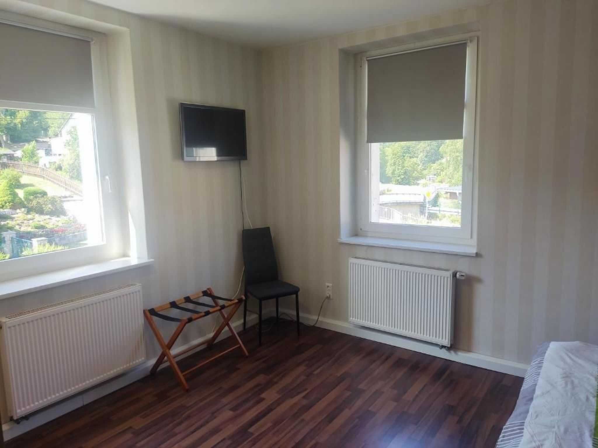Una habitación con dos ventanas, un radiador, una silla y un televisor en la pared.