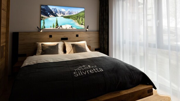 Un dormitorio con cama, televisor montado y ventana con cortinas translúcidas.