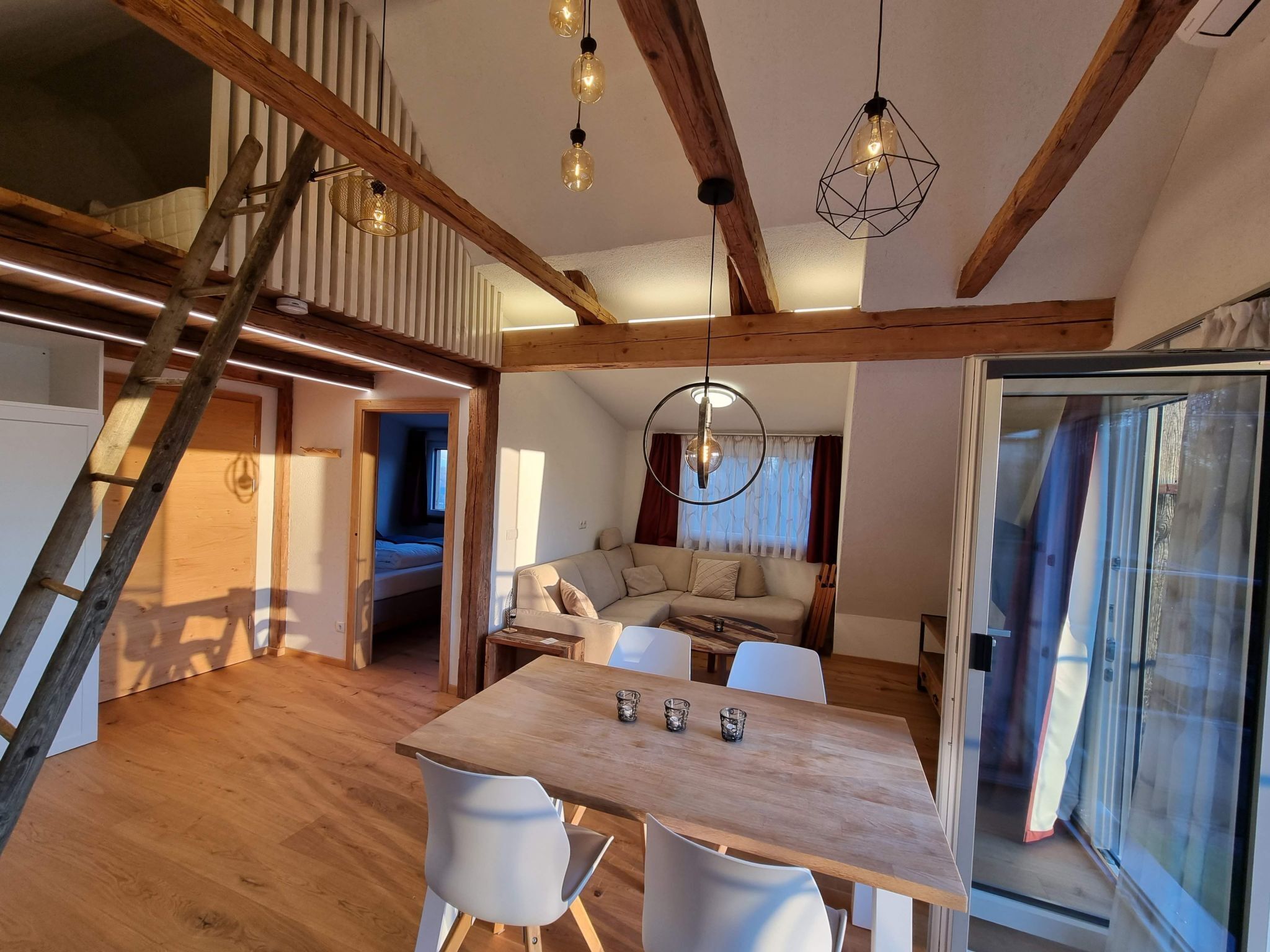 A modern loft-style room with exposed wooden beams, a wooden dining table with four chairs, a sofa, and a large window with curtains.