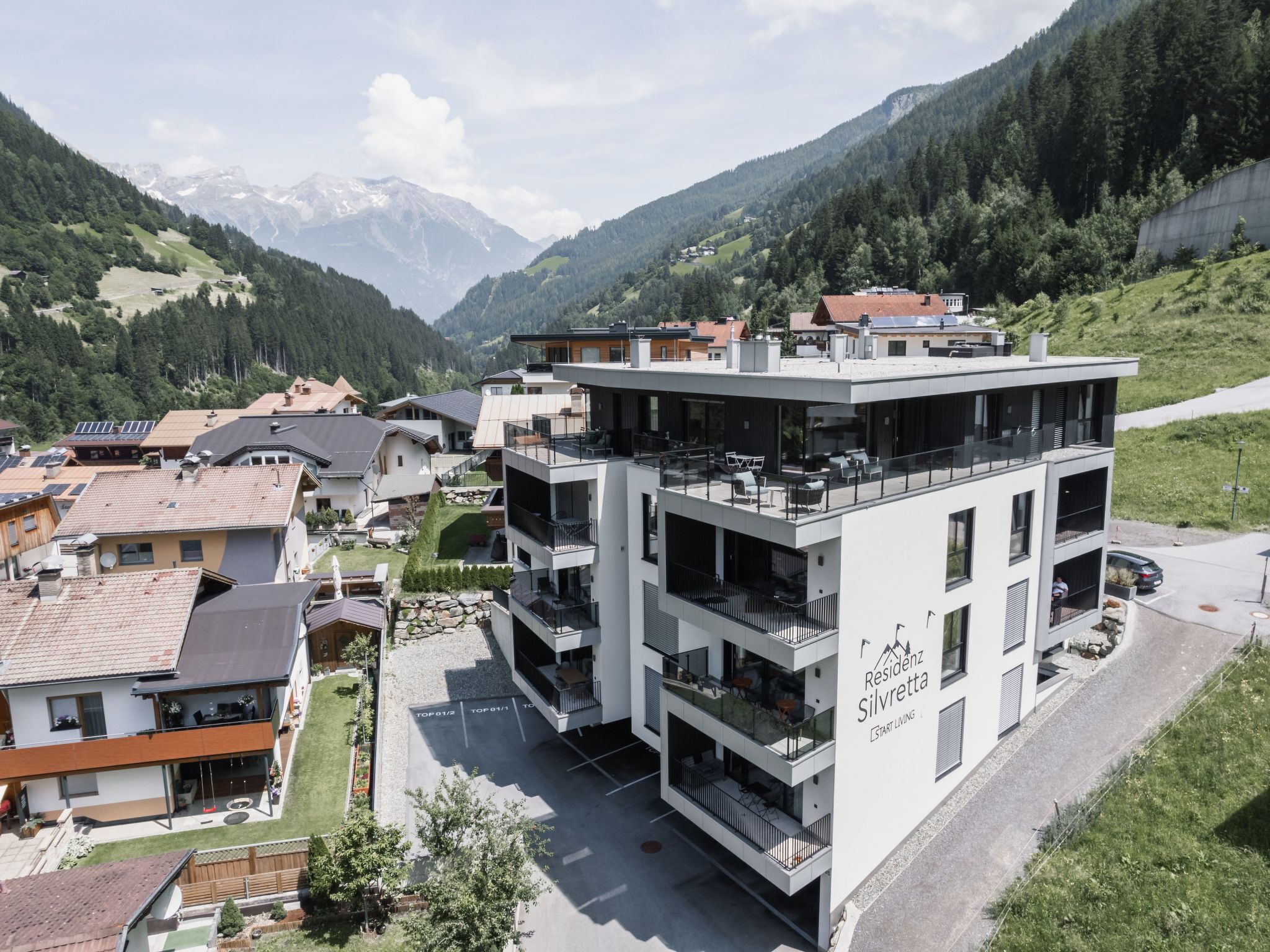 A modern residential building with multiple balconies, situated in a mountainous area with houses and greenery around.