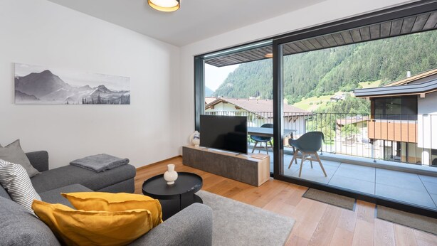 Ett modernt vardagsrum med en grå soffa, en gul prydnadskudde, ett svart soffbord och en platt-TV. Ett stort fönster ger utsikt över ett berg och en byggnad.