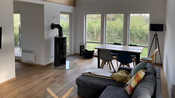 Uma sala de estar moderna com teto de madeira, lareira a lenha preta, mesa de jantar com cadeiras e grandes janelas com vista para um jardim.
