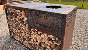 Een robuuste, buitenbarbecue met een metalen buitenkant en een houten binnenkant, gelegen in een grindgebied en omringd door een hek.