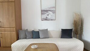 Un saló acollidor amb un armari de fusta, un sofà beix amb coixinets grisos i fosc, una taula de fusta petita amb un bol, i un quadre penjat al mur que representa un paisatge marí.