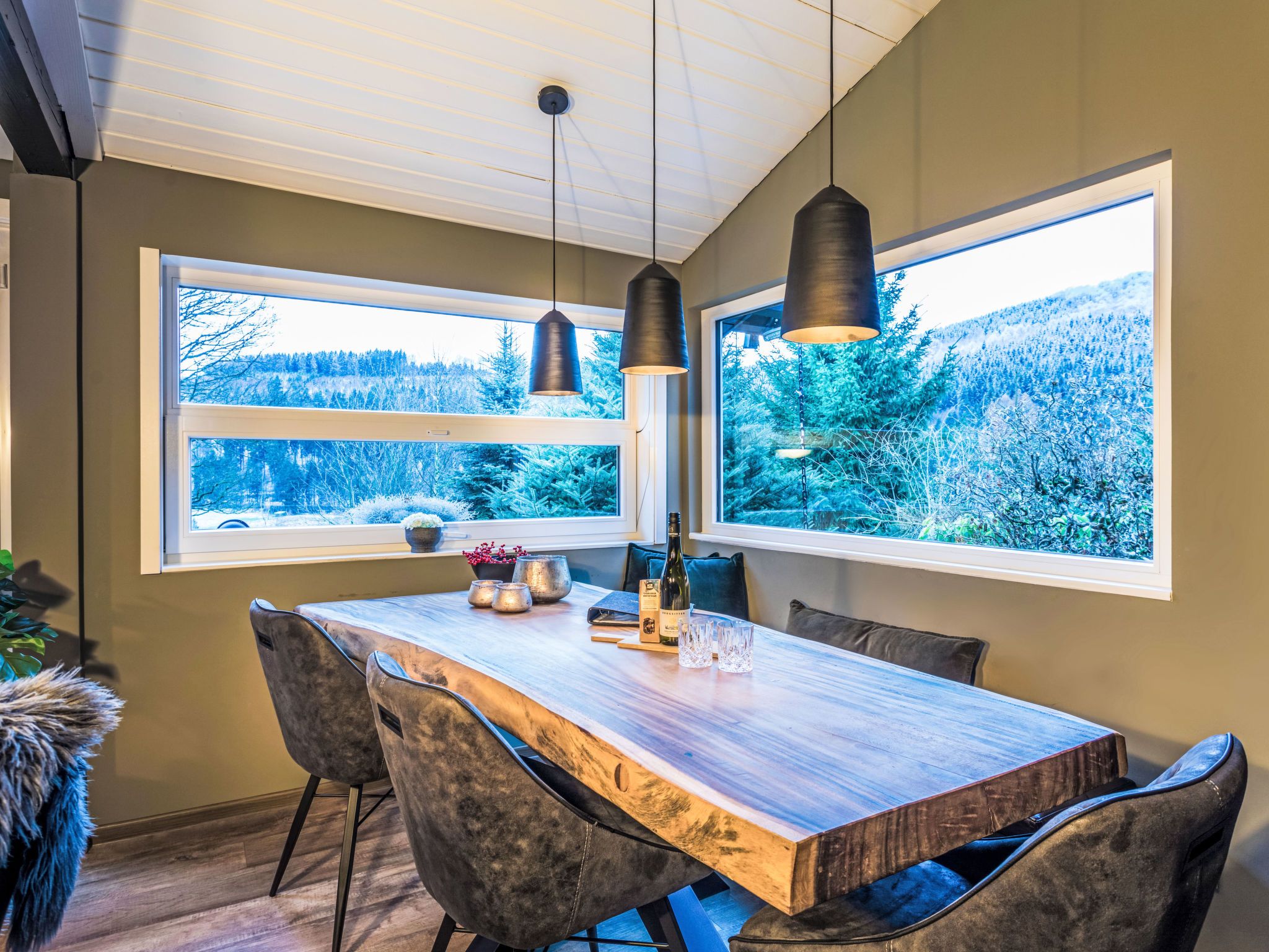 A dining area with a wooden table, chairs, pendant lights, and large windows offering a view of a forest.