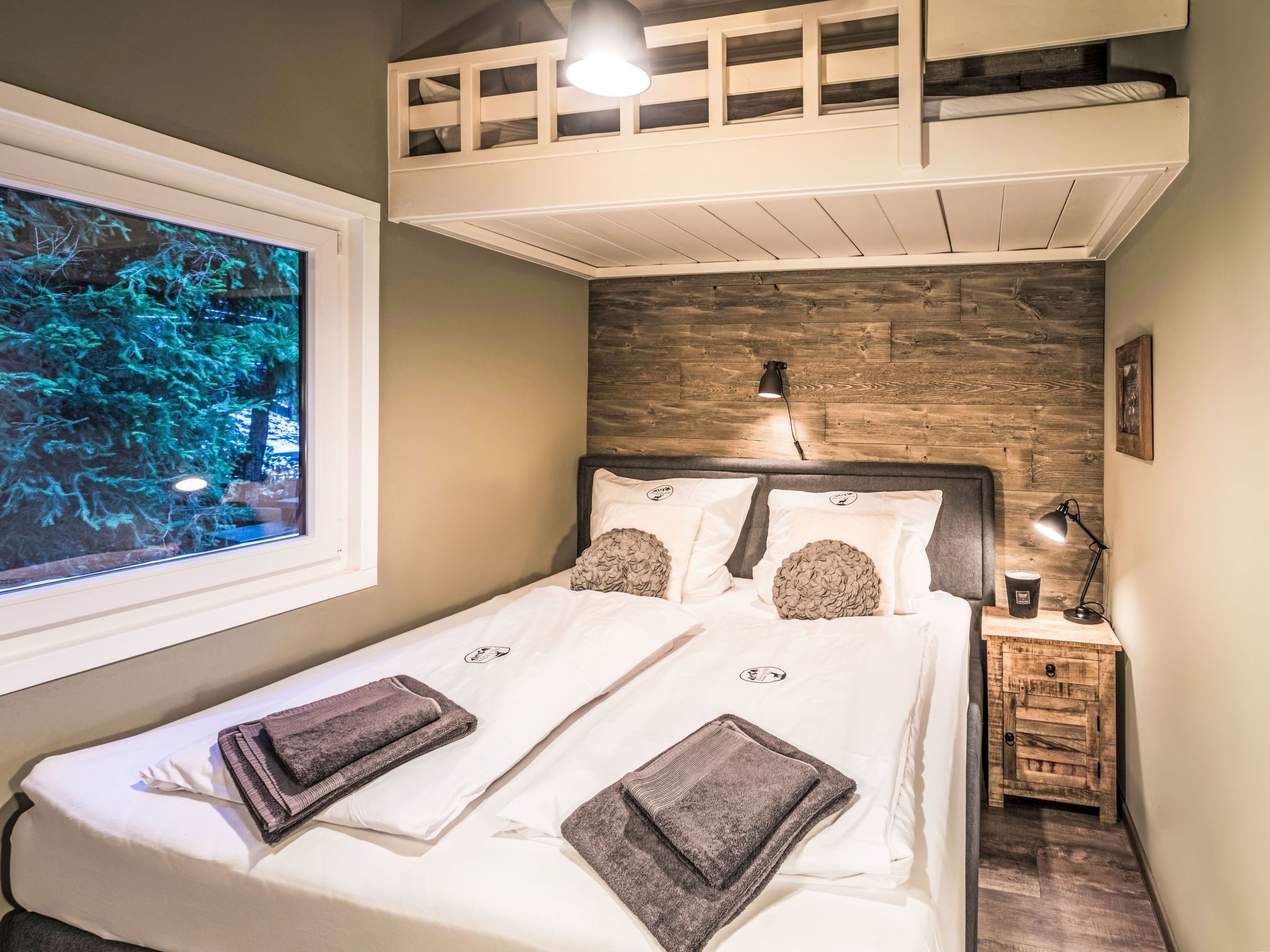 A bedroom with a bunk bed, a wooden headboard, a bedside table with a lamp, and a large window with a view of trees.